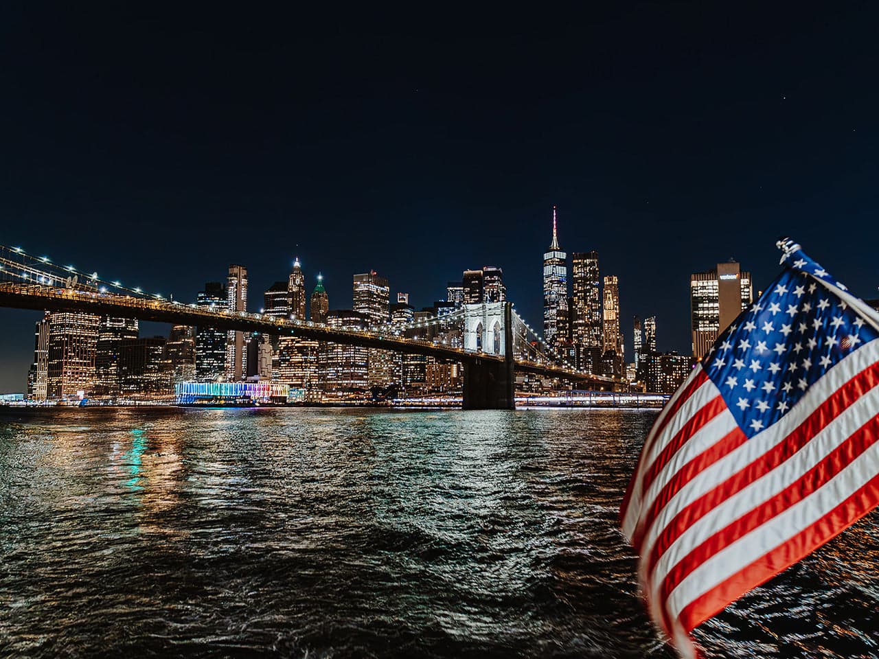 The Brooklyn Bridge and lower Manhattan lit at night, seen from the deck of a NYC Water Cruises vessel