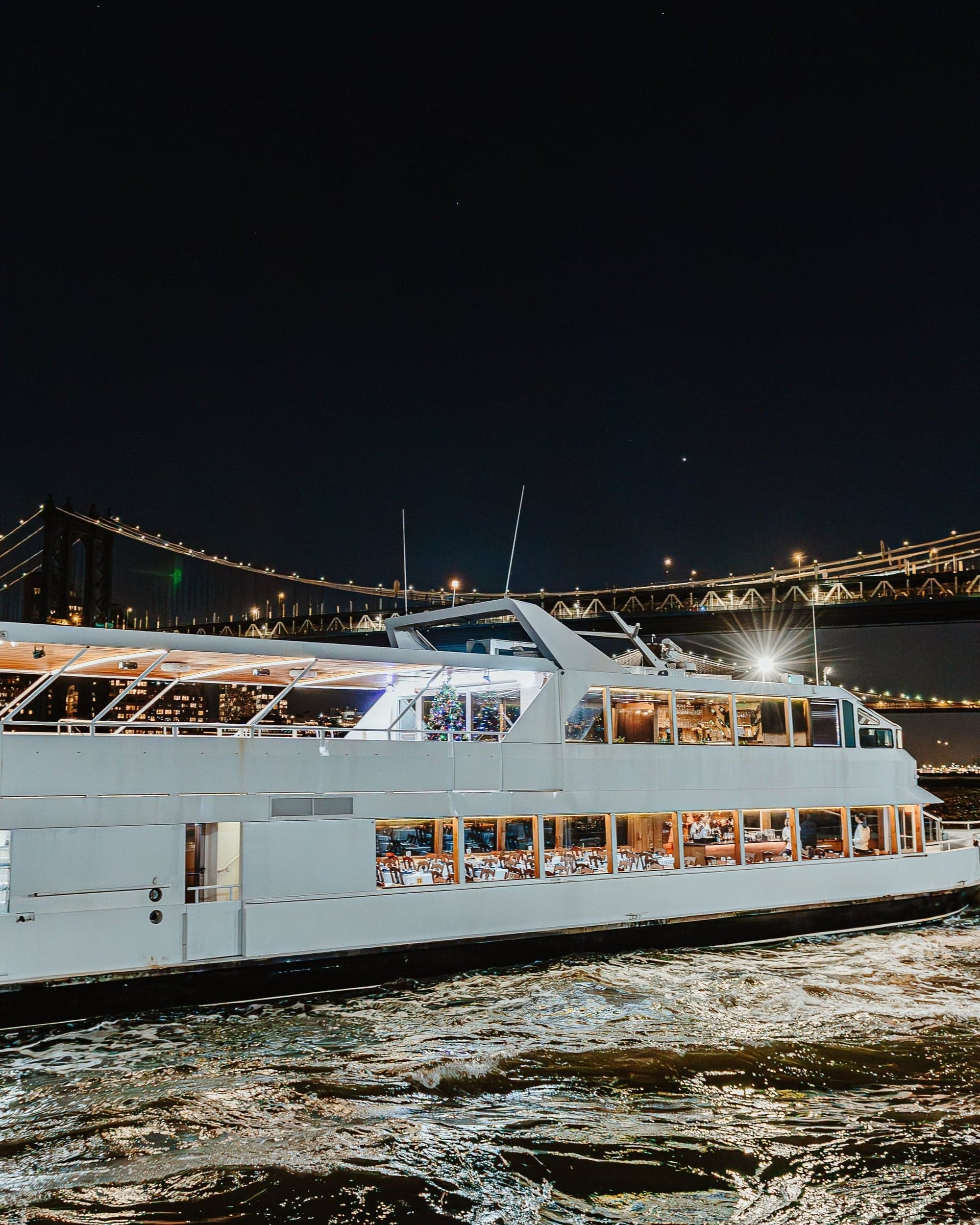 A NYC Water Cruises vessel passing beneath the Manhattan Bridge at night