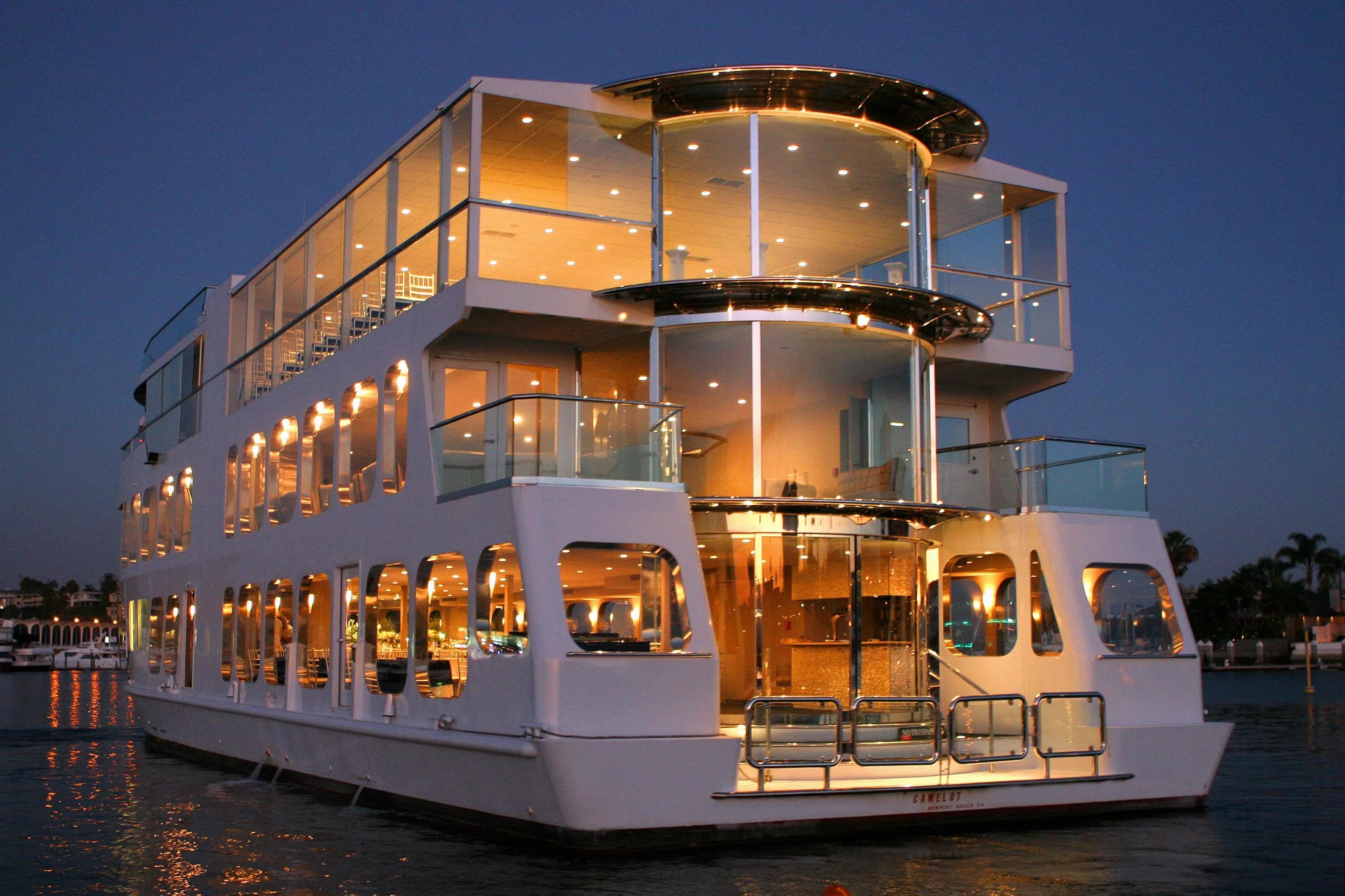 A large, multi-deck luxury yacht illuminated with warm lights at dusk. The sleek, modern vessel features expansive glass windows, multiple balconies, and polished metal finishes. It is anchored on a calm body of water during the Macy’s fireworks show, with a beautiful evening sky in the background. This is NYC Water Cruises Inc.'s July 4th: Eternity Dinner Cruise.