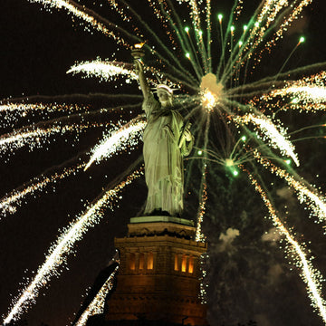 The Statue of Liberty is illuminated against a dark night sky with spectacular fireworks from the Macy’s fireworks show exploding in vibrant colors behind it, creating a radiant display of streaking lights and bursts. The statue stands majestically, holding her torch high. For an unforgettable experience viewing this stunning sight, join the July 4th: Eternity Dinner Cruise by NYC Water Cruises Inc.
